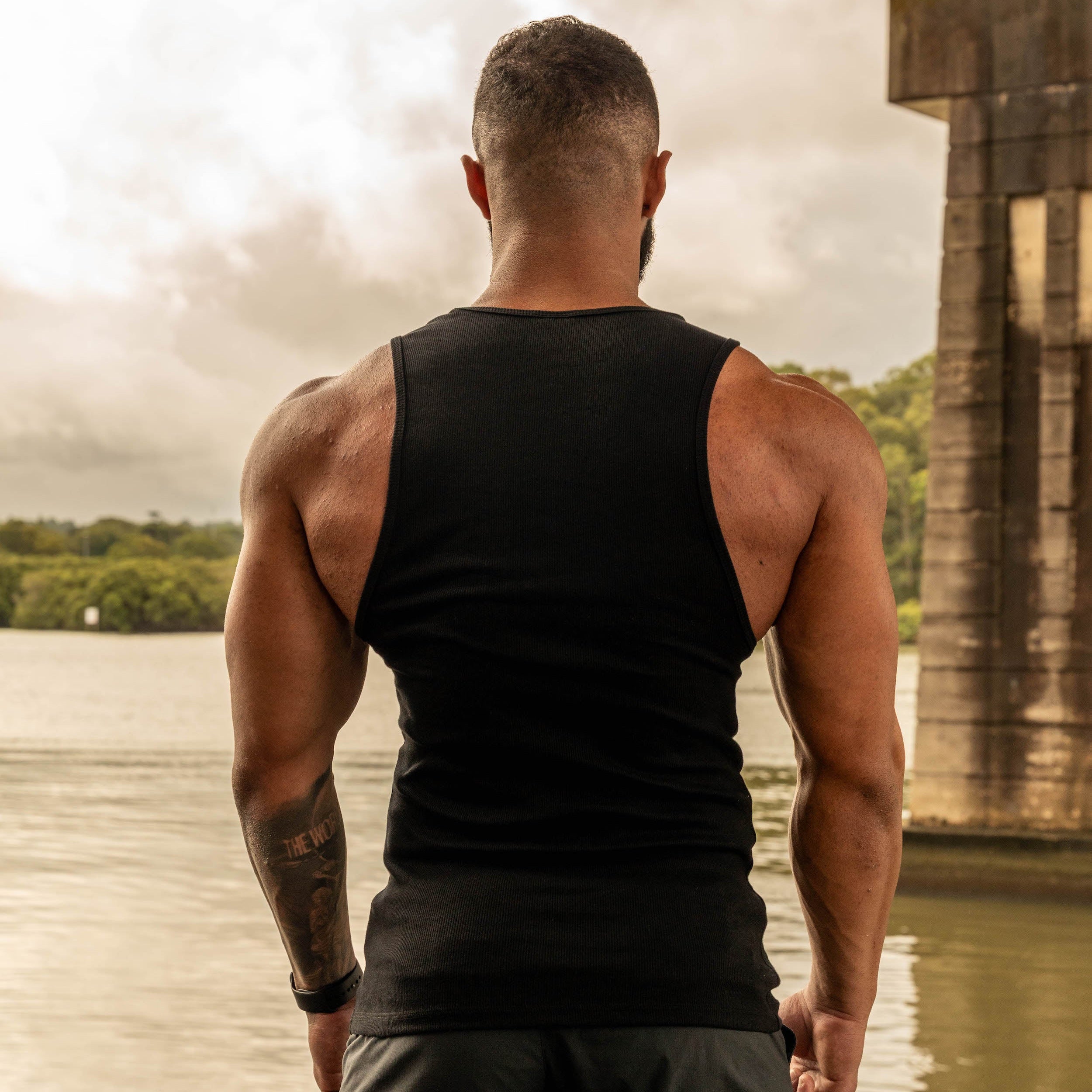 back pose showing muscular physique in black tank top by water